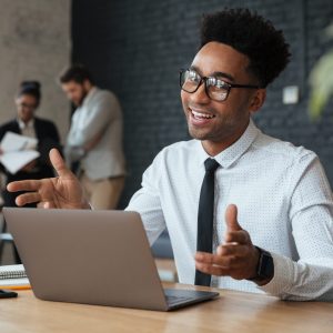 Photo of happy young african businessman sitting indoors using laptop computer. Looking camera.